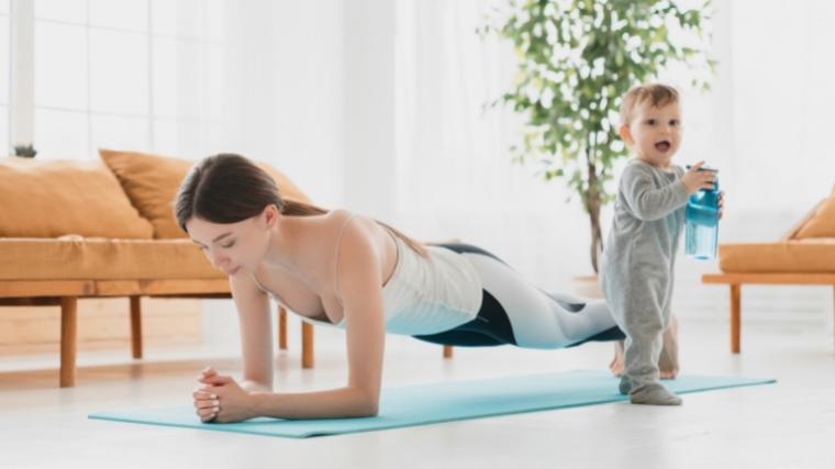 A woman performing a plank with a small child standing next to her holding a bottle