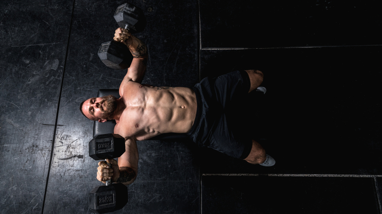 Muscular man in dark gym lying on bench while pressing dumbbells