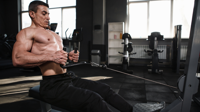 Shirtless muscular man in dark gym performing seated row exercise