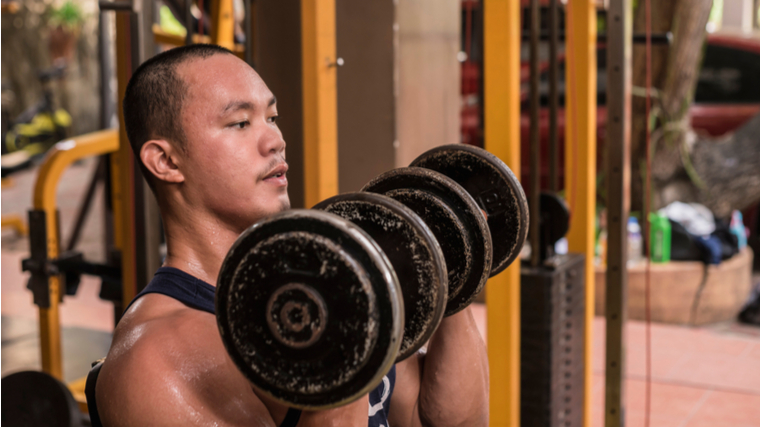 Man sitting in gym holding dumbbells at shoulders
