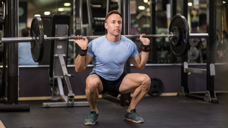 Man in blue t-shirt wearing wrist wraps performing a back squat in a loaded barbell