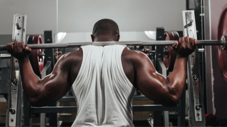 Man in white sleeveless t-shit squatting with a loaded barbell wideness his back