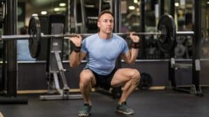 Man in blue t-shirt wearing wrist wraps performing a back squat in a loaded barbell