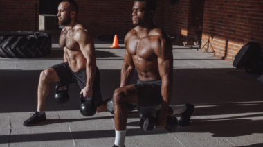two muscular people in dark gym performing dumbbell lunge