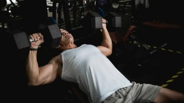 person in white tank top presses two dumbbells over his chest on an incline.