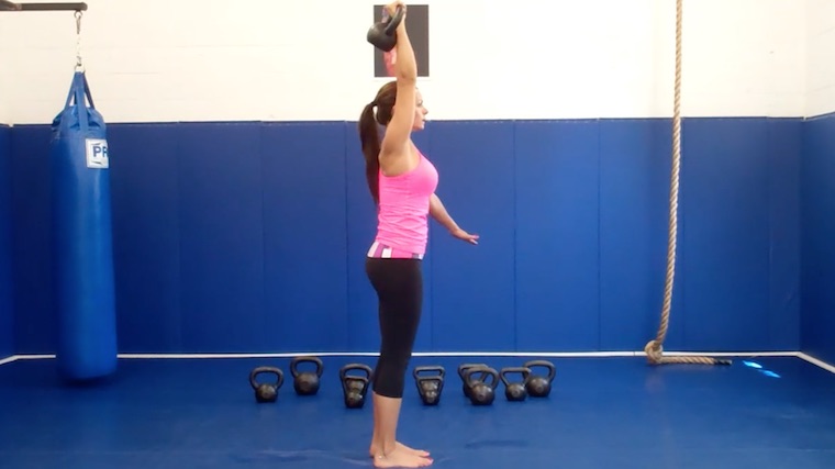 Long-haired person in gym standing with kettlebell overhead