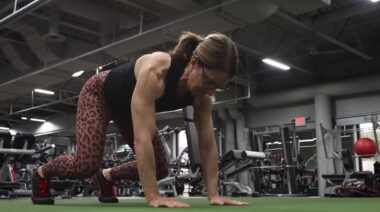 Long-haired person wearing glasses in gym on ground performing bear plank.