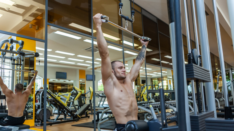 Shirtless muscular person in gym doing cable pulldown exercise