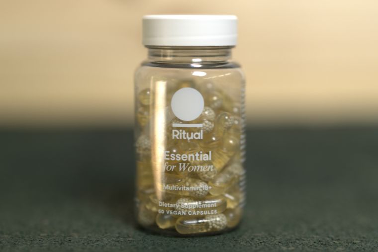 A bottle of the Ritual Essential Women's Multivitamin 18+ on a black surface with a wood background