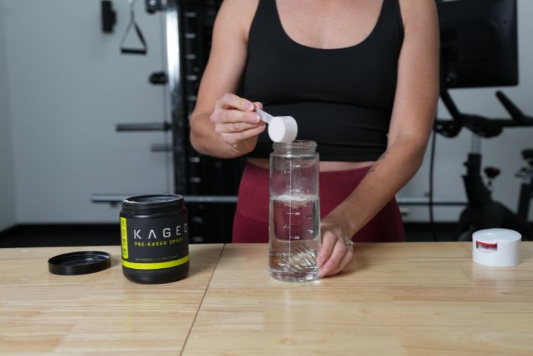 A woman pouring a scoop of Kagged Pre-Kagged Sport into a water bottle