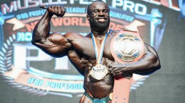 Samson Dauda flexes his right biceps while holding the title belt for winning the 2023 Romania Muscle Fest Pro.